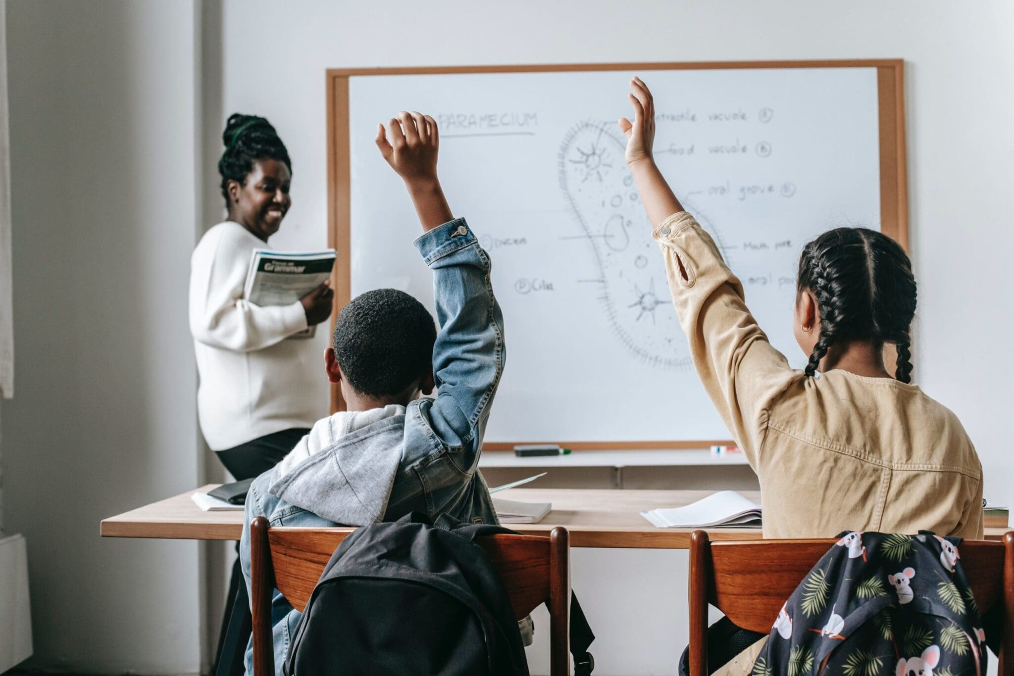 Classroom with two students raising their hand, facing a smiling teacher at a while board