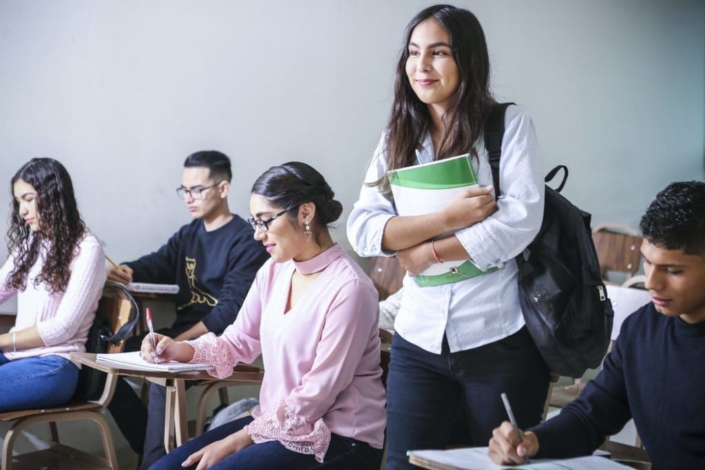 Young woman standing in classroom with books while other students sit at desks