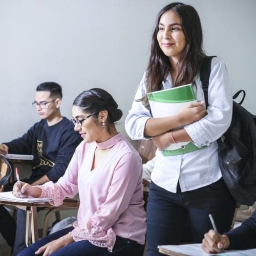 Young woman standing in classroom with books while other students sit at desks