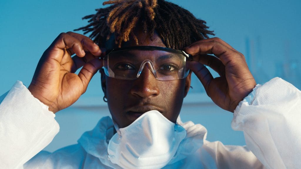 Young Black man in lab coat and mask pulled down adjusting goggles against a blue backdrop.