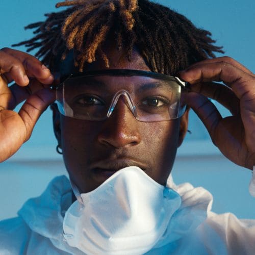 Young Black man in lab coat and mask pulled down adjusting goggles against a blue backdrop.