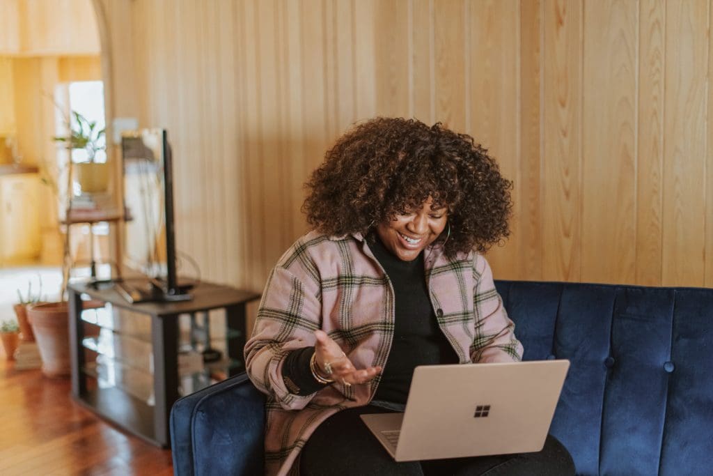 Black woman with natural hair sitting on a couch with her laptop. She is speaking to someone on the screen.