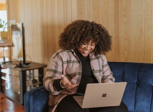 Black woman with natural hair sitting on a couch with her laptop. She is speaking to someone on the screen.