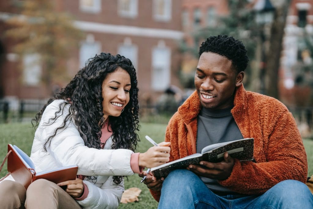 Two students sitting on the grass at college, smiling and looking at a notebook