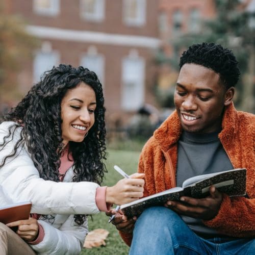 Two students sitting on the grass at college, smiling and looking at a notebook