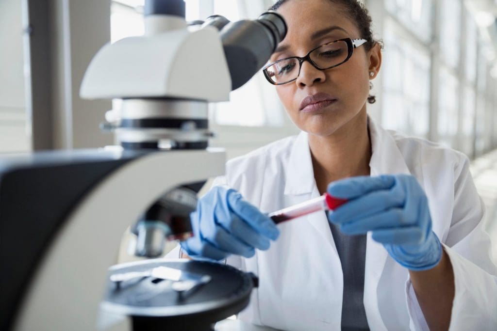 Black woman with lab coat and gloves working with microscope.