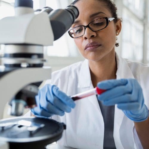 Black woman with lab coat and gloves working with microscope.