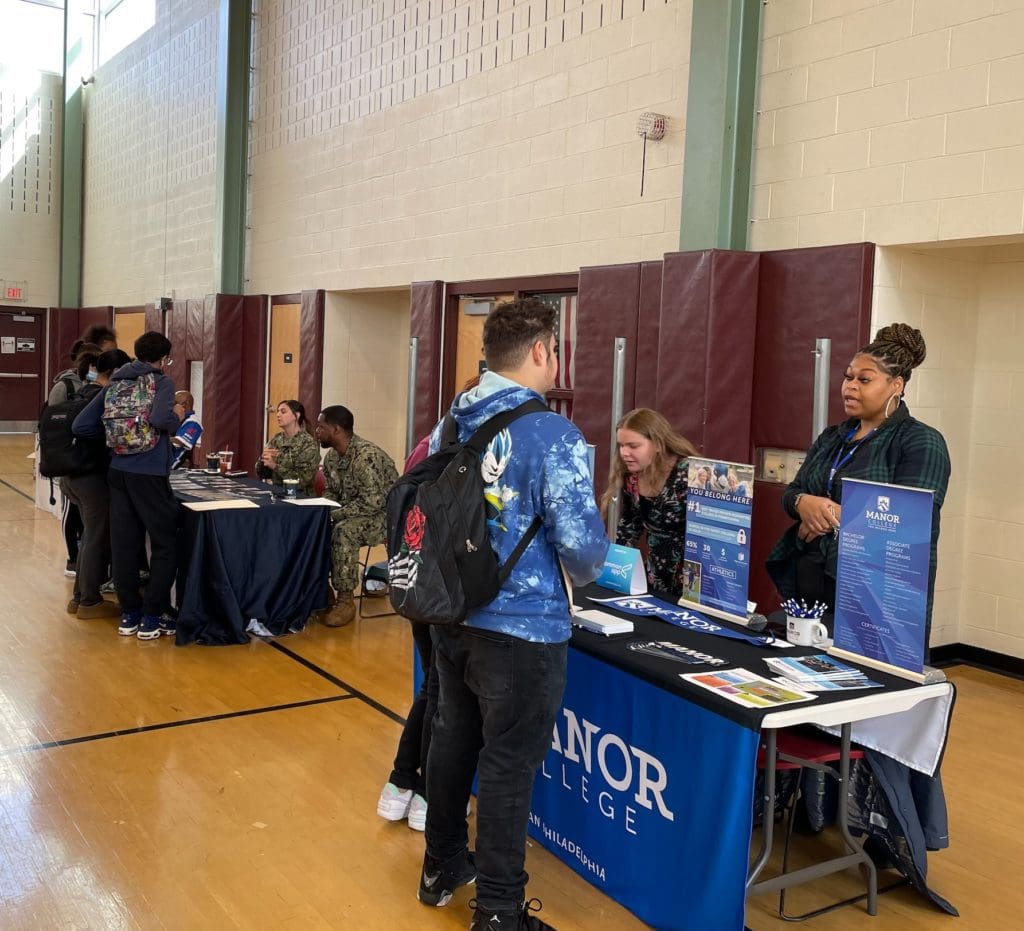 Student and college representative talking over a display table in a high school gym.