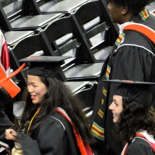 Two women in caps and gowns moving through an auditorium and smiling. 