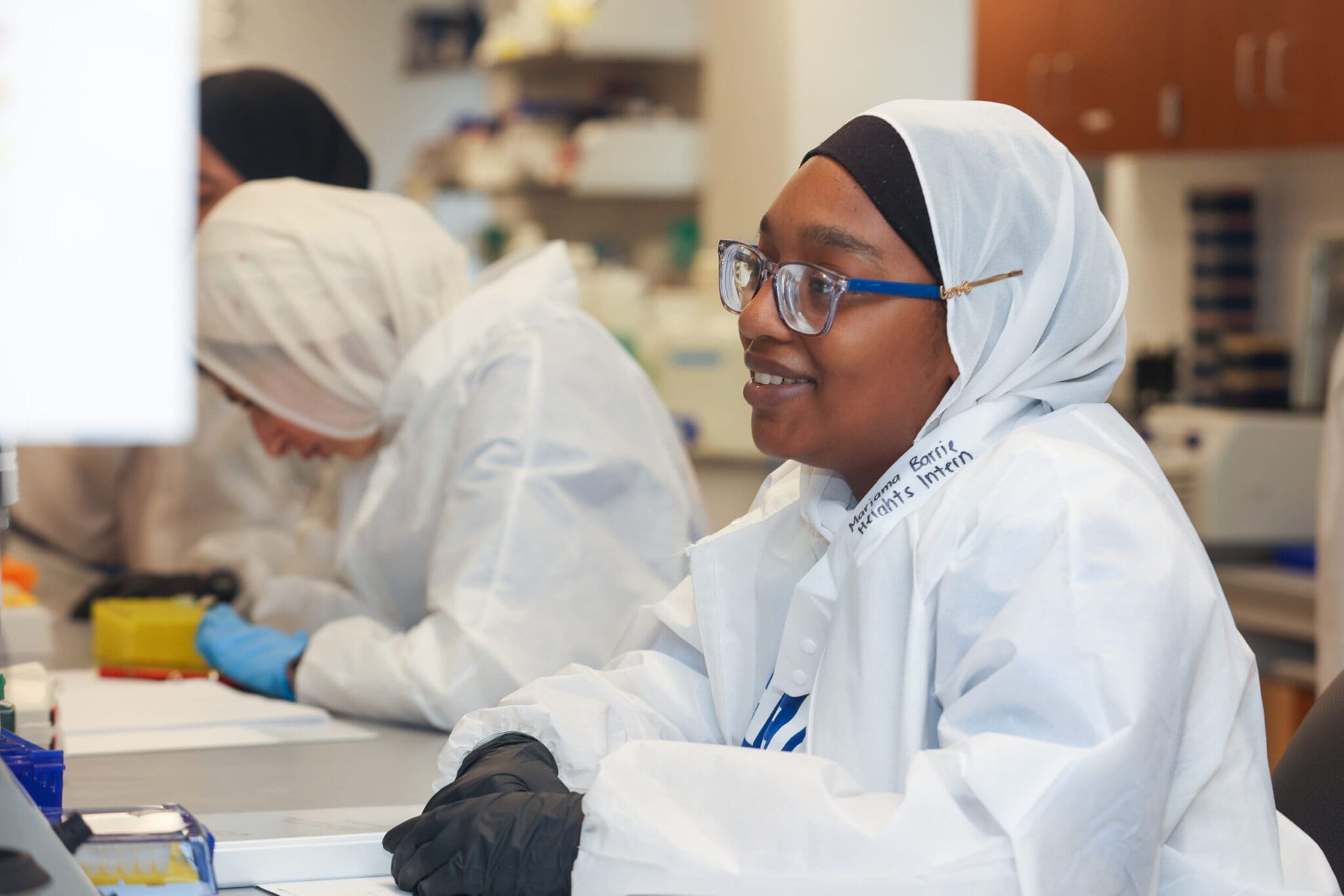 Black woman with lab coat and gloves working with microscope.