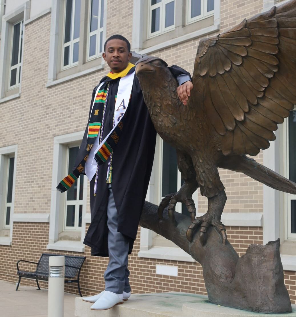 Student in graduation gown and stole posing with St Joseph's University hawk sculpture.